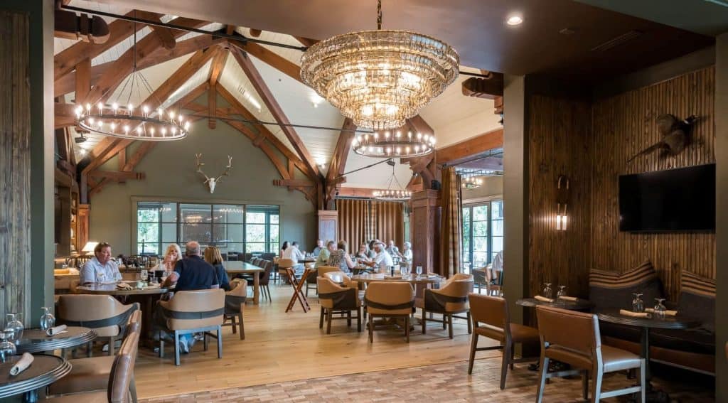 Spacious restaurant with wooden beams, large chandeliers, and several groups of people dining at round tables during the day.