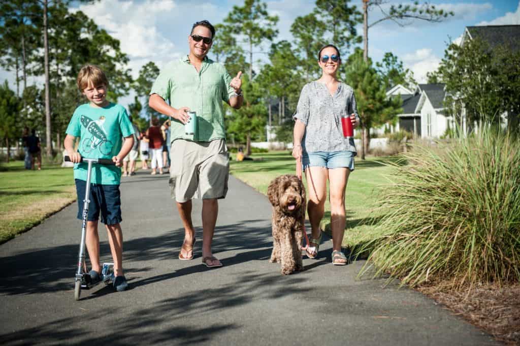 A boy rides a scooter, a man walks and gives a thumbs up, and a woman walks a dog on a sunny path with houses and trees in the background.
