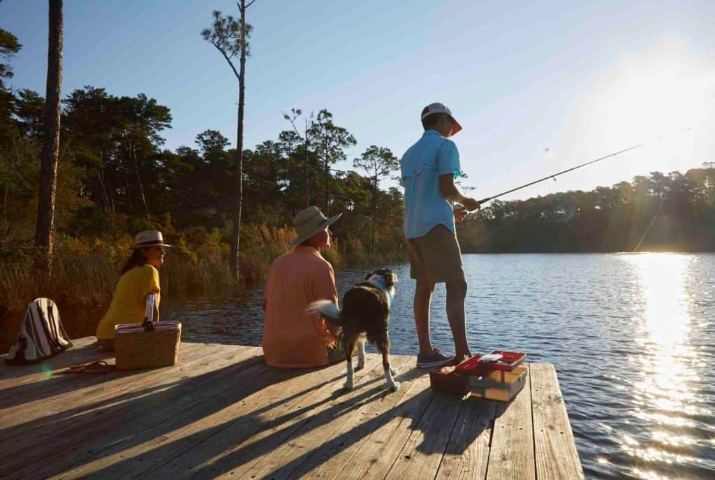Three people and a dog sit and stand on a wooden dock by a lake, fishing in the late afternoon sunlight. A picnic basket and model boat are on the dock.