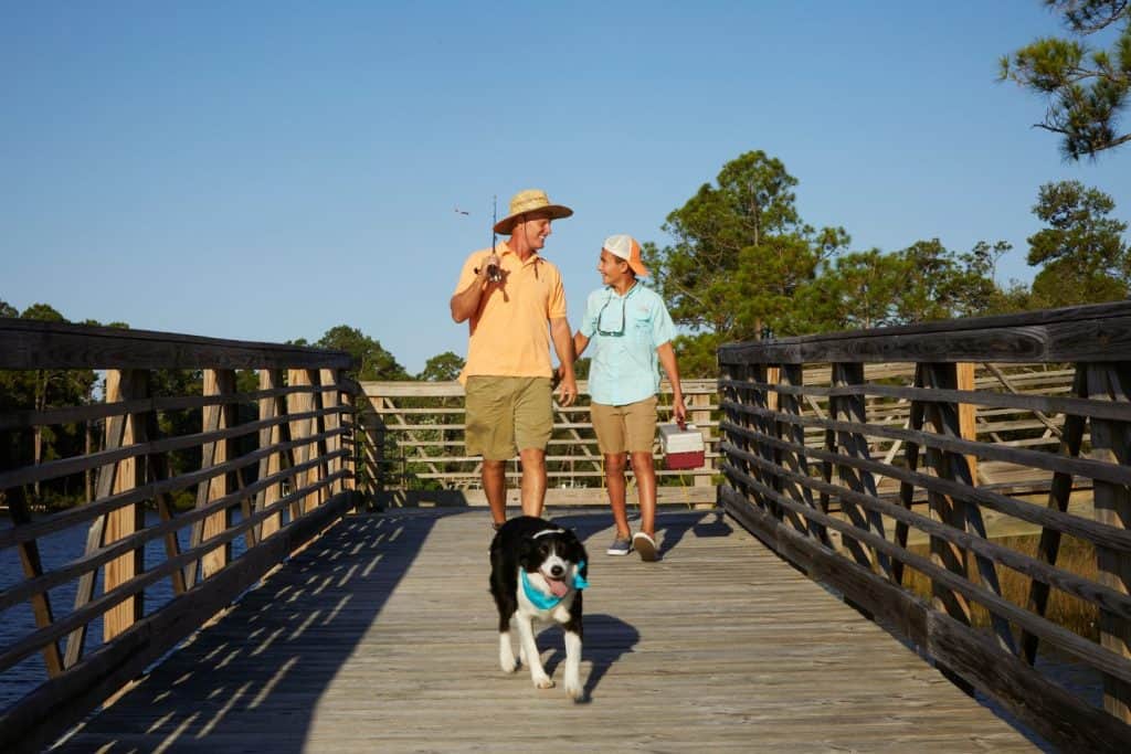 Two people walk on a wooden bridge with a fishing rod and cooler, accompanied by a black and white dog. Trees and blue sky are visible in the background.