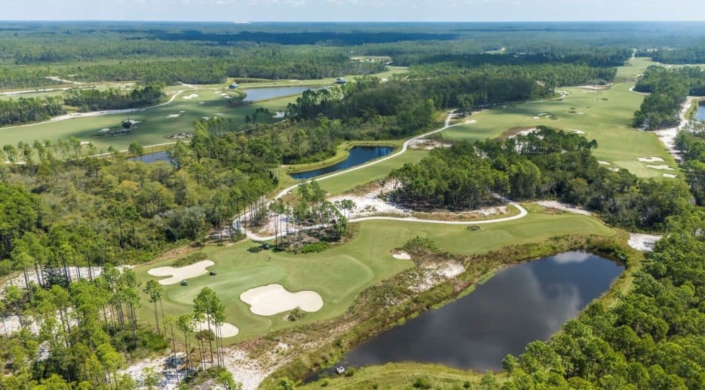 Aerial view of a golf course with multiple sand bunkers, ponds, winding paths, and surrounding forested areas under a clear sky.
