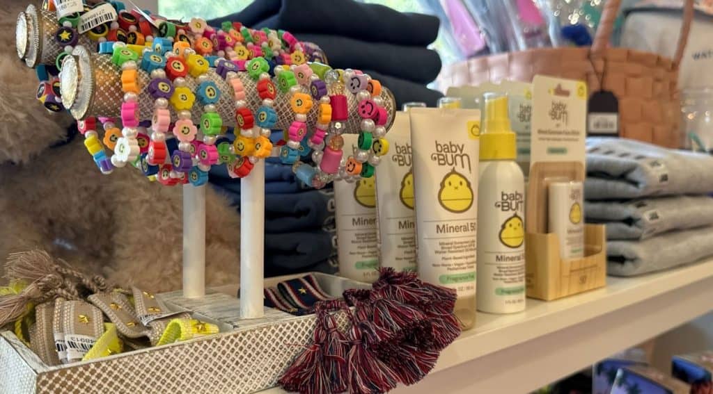 Colorful beaded bracelets, Baby Bum skincare products, folded clothing, and woven tassel keychains displayed on a store shelf.