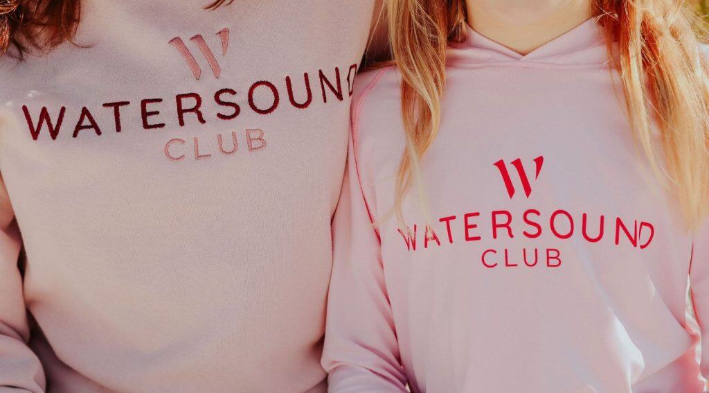 Two people wearing light pink "Watersound Club" sweatshirts with red lettering, shown from shoulders to mid-torso.