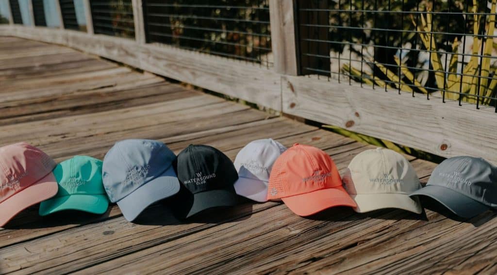 Eight baseball caps in various colors are lined up on a wooden boardwalk next to a fence on a sunny day.