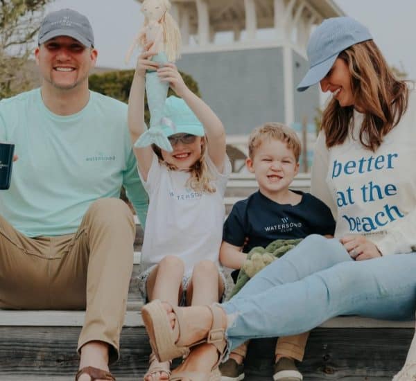 A family of four sits on outdoor wooden steps, smiling. The father holds a mug, the mother wears a "better at the beach" sweatshirt, and the children hold toys.