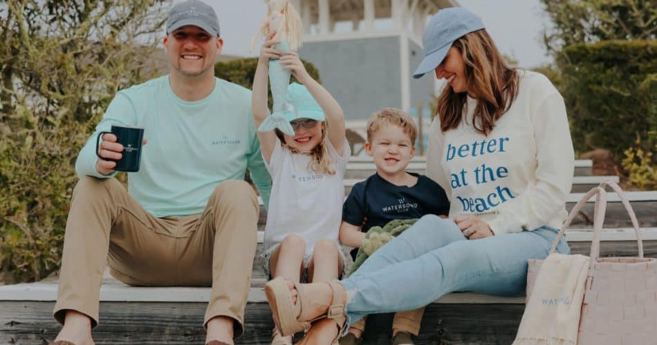 A family of four sits on outdoor wooden steps, smiling. The father holds a mug, the mother wears a "better at the beach" sweatshirt, and the children hold toys.