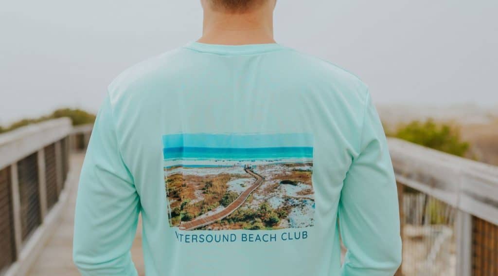 Person wearing a light blue long-sleeve shirt with a beach scene and the text "WATERSOUND BEACH CLUB" printed on the back, standing on a boardwalk.