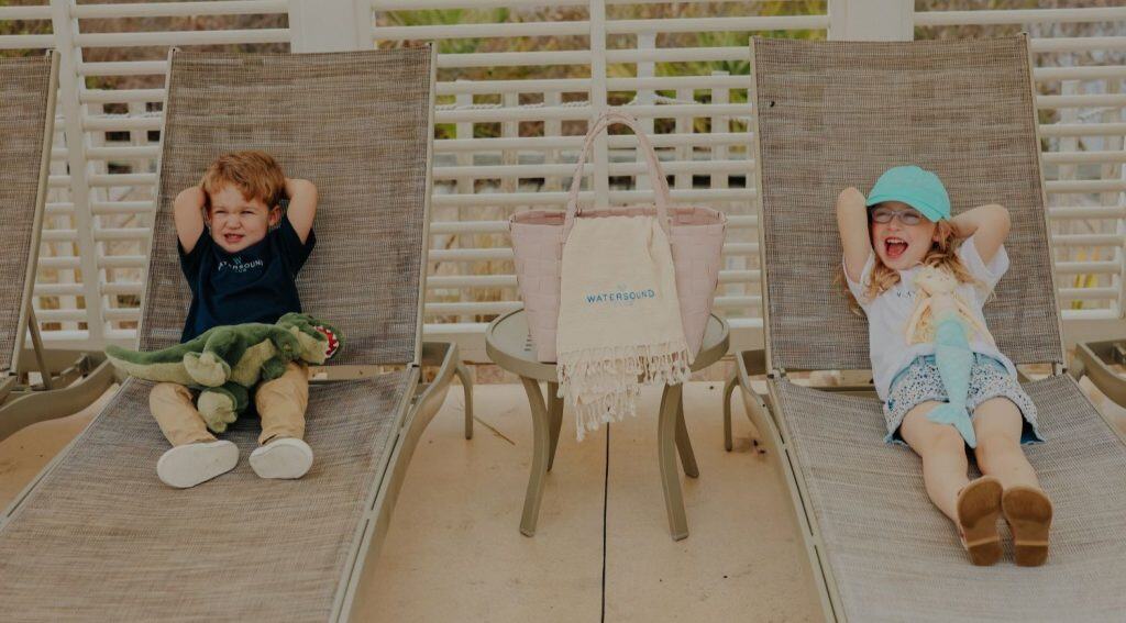 Two children relax on lounge chairs by a pool, with a tote bag and towel on a small table between them. The boy holds a stuffed dinosaur, and the girl wears a mint cap and sunglasses.