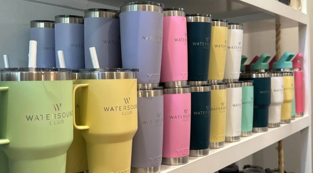Several pastel-colored and metallic tumblers with "Watersound Club" branding are neatly arranged on white shelves.