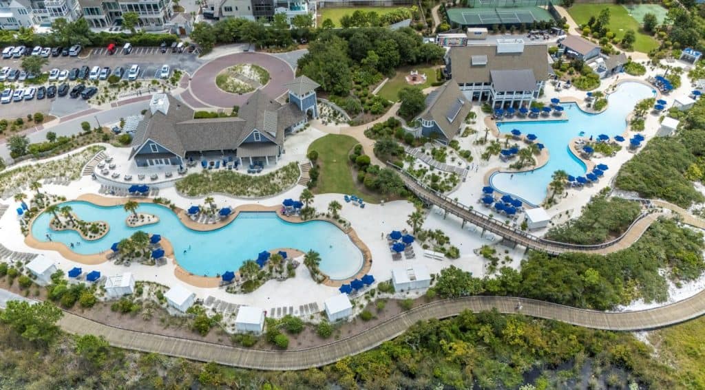 Aerial view of a resort with two large outdoor pools, blue lounge chairs, cabanas, several buildings, parking lot, and landscaped grounds surrounded by trees.