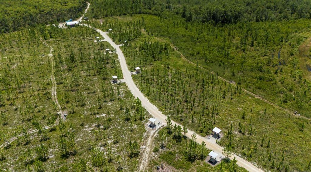 Aerial view of a dirt road winding through a green, sparsely forested area with small white structures scattered along the path.