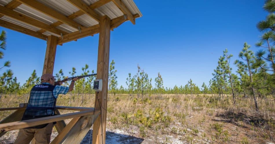 A person in a shooting vest aims a shotgun from a wooden platform at a clay target in a grassy, tree-lined outdoor shooting range under a clear blue sky.