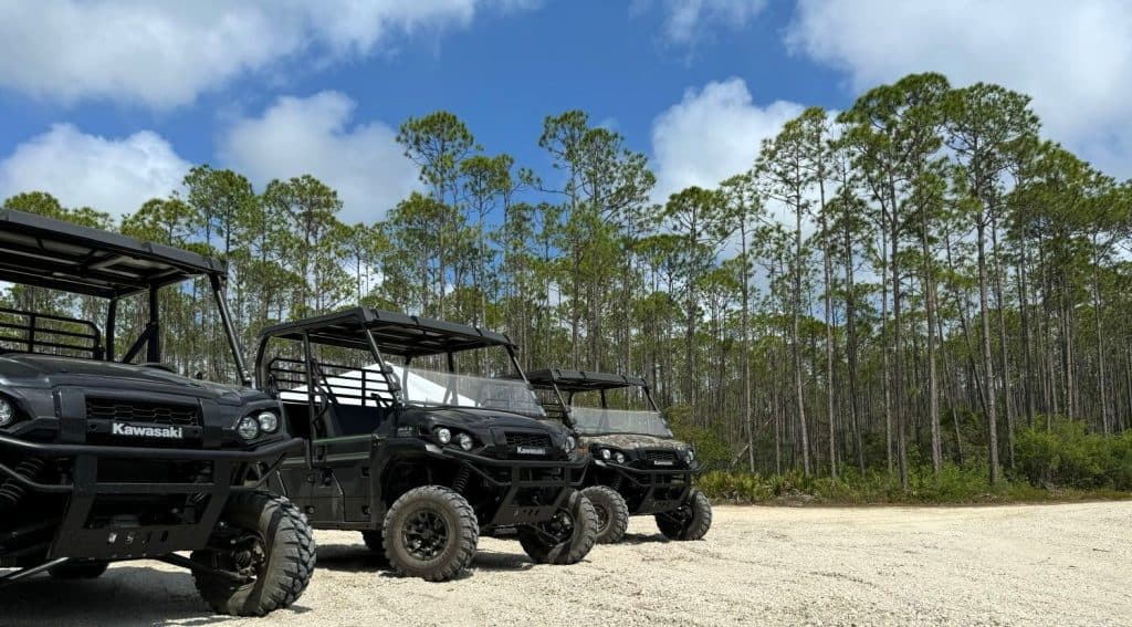 Three Kawasaki utility vehicles are parked on a sandy dirt lot, with tall pine trees and a blue sky with clouds in the background.