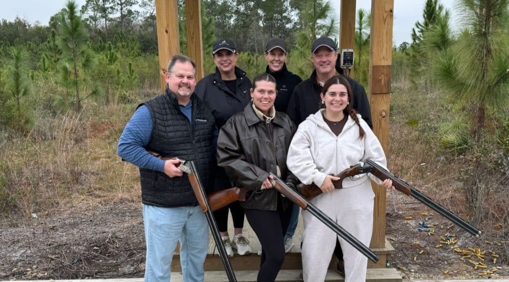 Six people pose outdoors at a shooting range, holding shotguns, standing on a wooden platform surrounded by trees. Spent shell casings are visible on the ground.